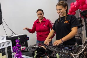 Chan in a fuscia adidas sweater behind a smiling teen in a black t shirt at a DJ mixing board